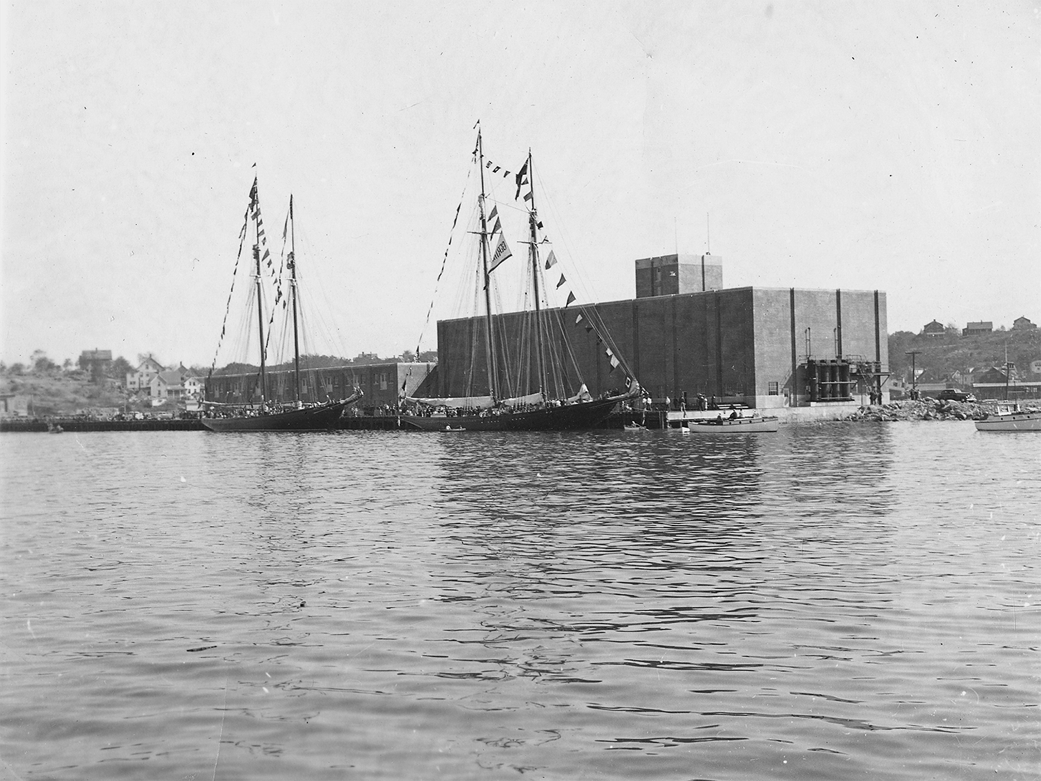 A historical waterfront scene with two large sailing vessels decorated with flags docked near an industrial-looking building