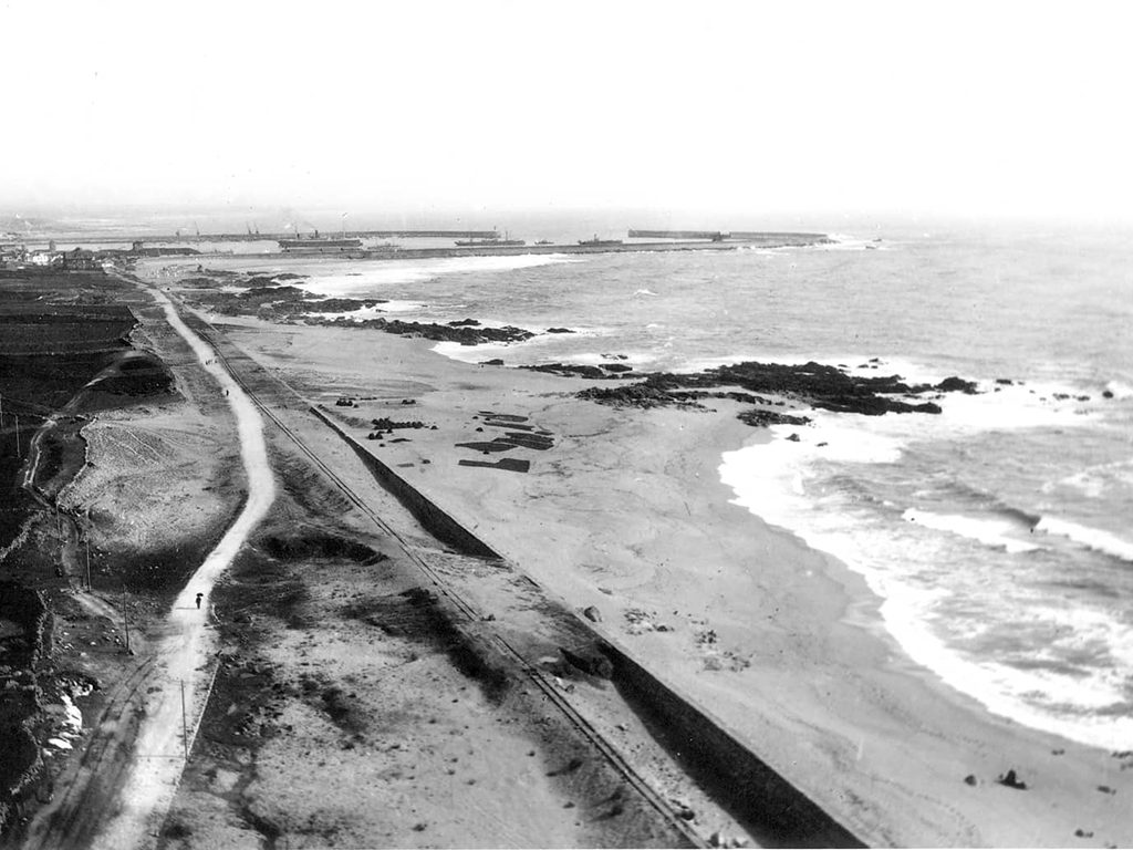 A wall, a railway and a path run parallel to the sea, extending into the distance to a harbour with ships.