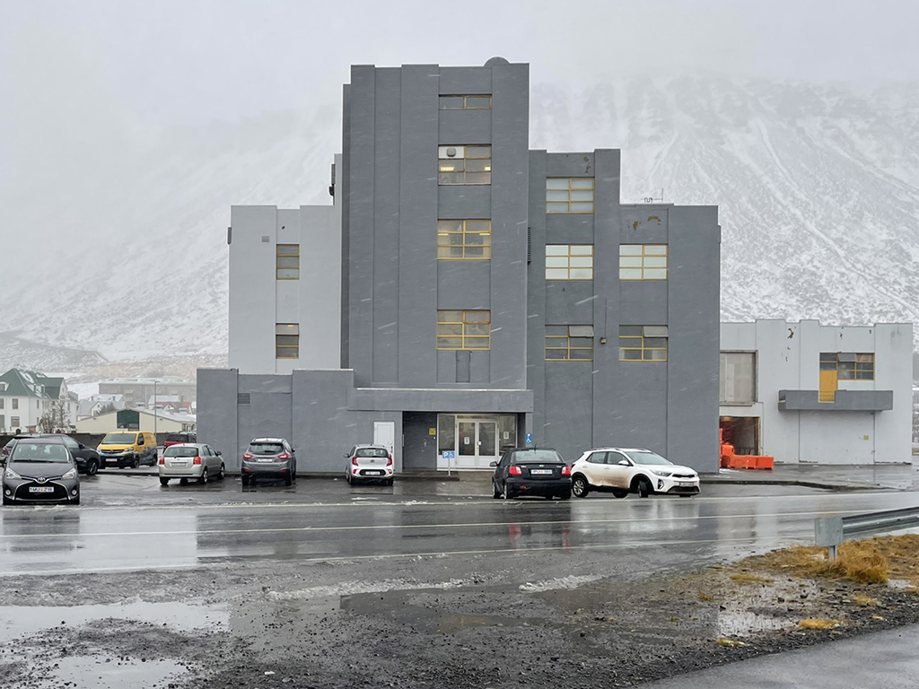 A prominent grey and white building with parking lot at the front. The asphalt is wet, picturing a rainy day.