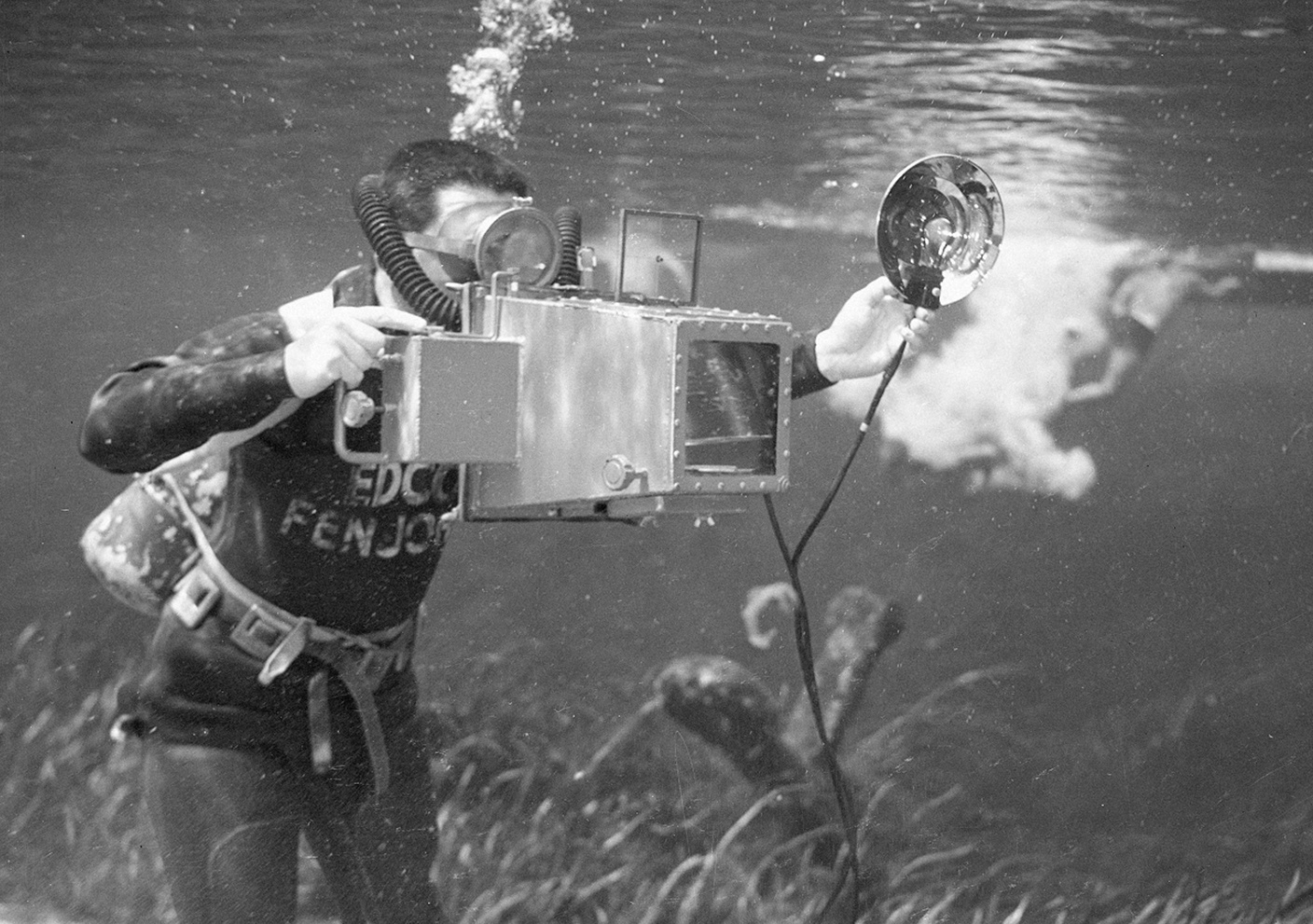 A scuba diver underwater is using a vintage camera with a large housing and an external flash.