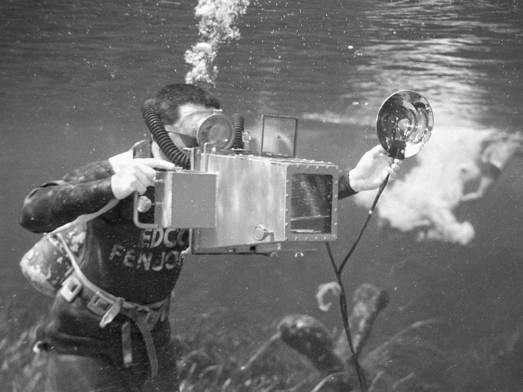 A scuba diver is underwater operating a large, vintage underwater camera with a flash attachment.