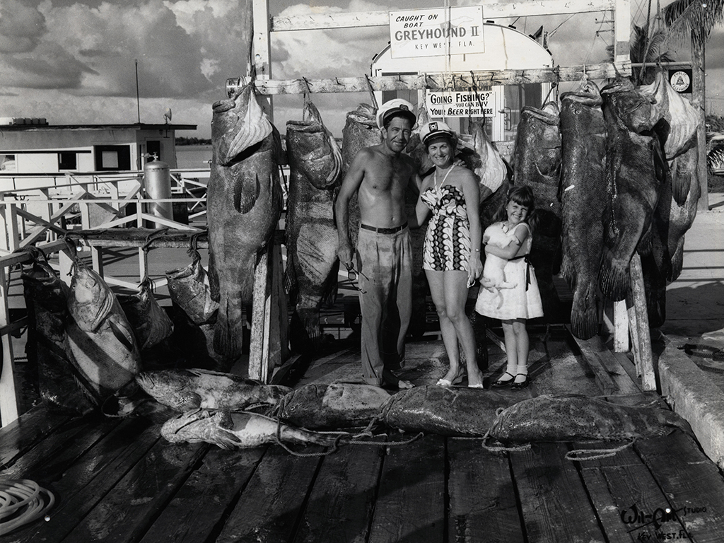 A fishing pear, portraying two adults and a child smiling next to massive groupers hanging and laying on the floor.